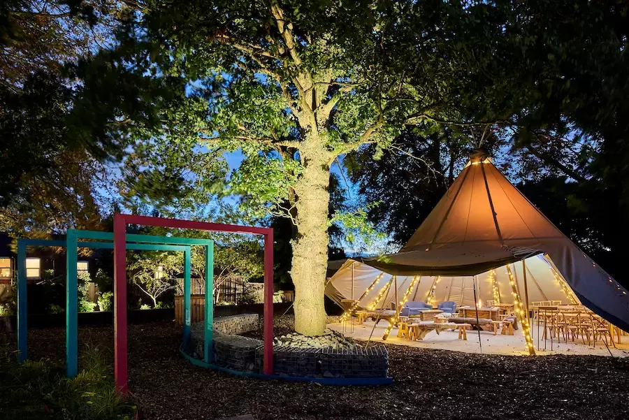 Luxurious Teepee tent furnished for weddng celebrations in landscaped garden with Oak tree and colourful arbours beautifully illuminated and lighting around the borders.
