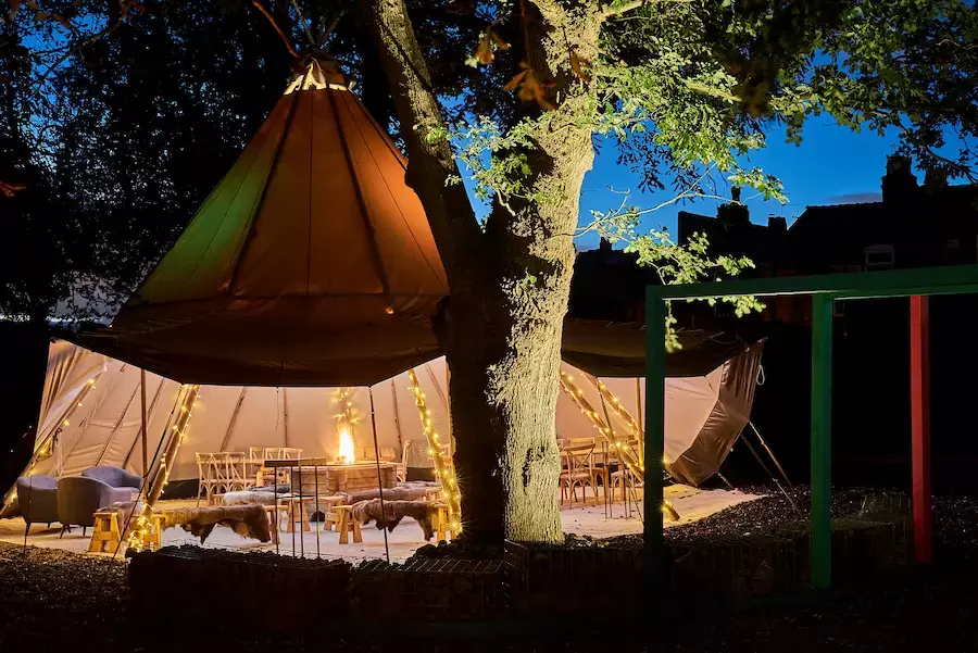 Oak tree and colourful arbours beautifully illuminated in large garden with luxurious teepee tent lavishly furnished for weddng celebrations.