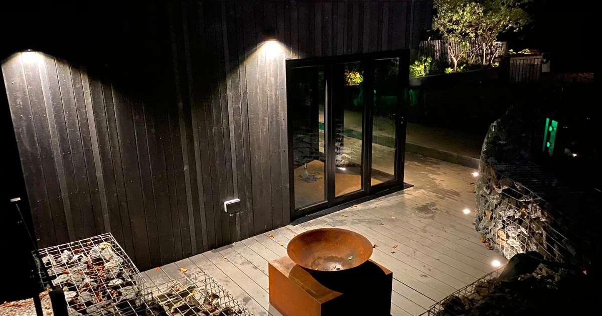 12v outdoor garden lighting installation completed in Harborne Birmingham. 230v outdoor wall lights illuminating garden studio walls and low voltage outdoor decking lights illuminating up gabion walled seating around sunken patio space with firebowl - Oakland Group, Outdoor Garden Lighting Design and Installation Services.