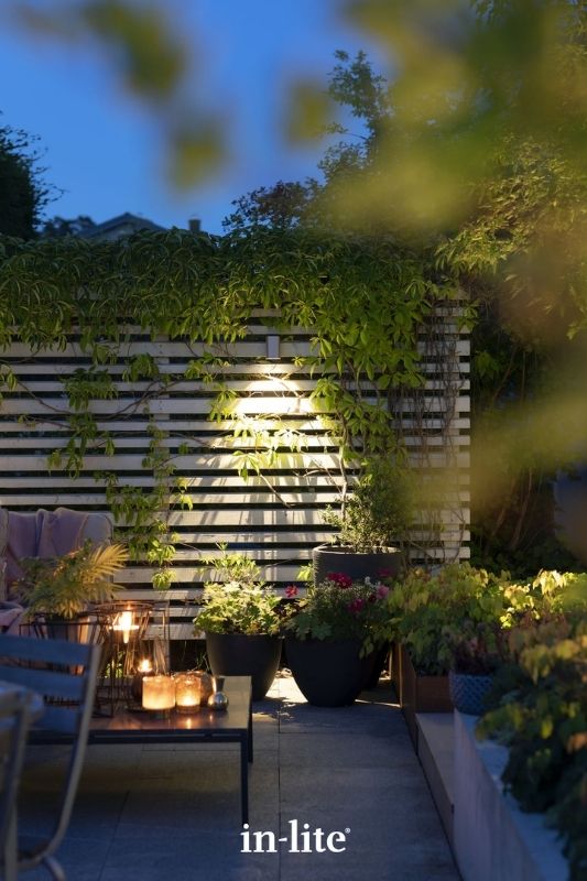 in-lite ACE DOWN 12v outdoor wall light, mounted on a slatted fence, illuminating low level planters positioned along patio border, with a targeted beam of light down to the ground. Low voltage outdoor garden lights, atmospheric lighting for ambiance.