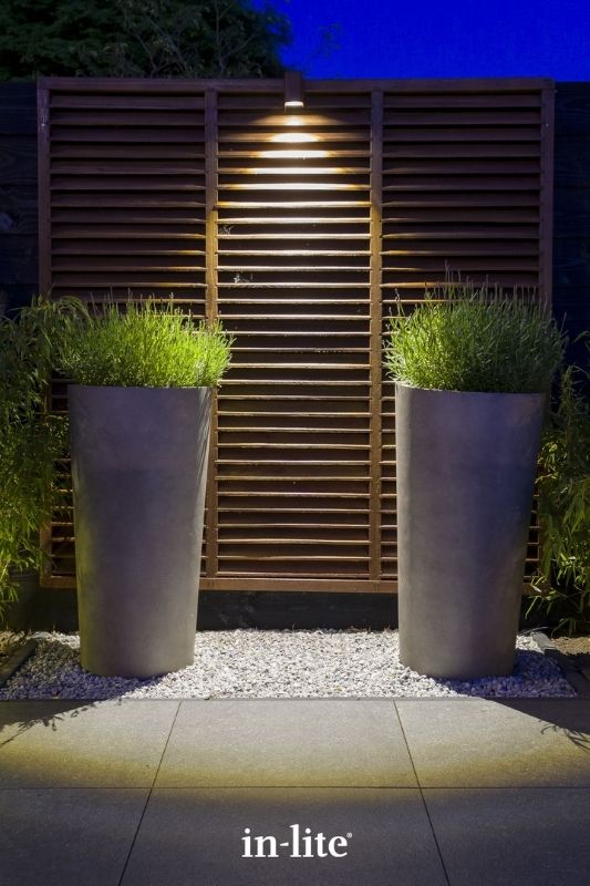 in-lite ACE DOWN CORTEN 12v outdoor wall light, mounted on a fence, illuminating tall planters positioned along patio border, with a targeted beam of downwards light to the ground. Low voltage outdoor garden lights, atmospheric lighting for ambiance.