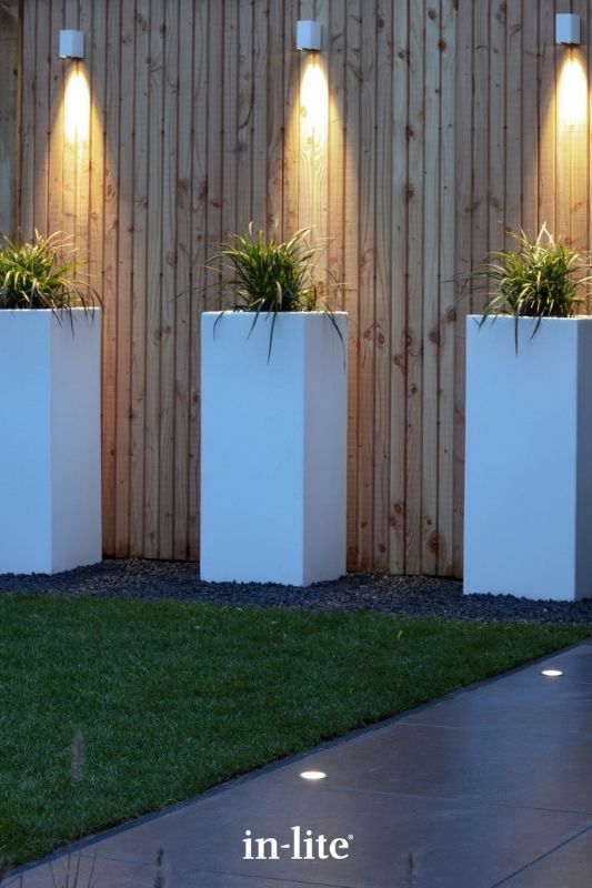 in-lite ACE DOWN WHITE 12v outdoor wall lights, installed along fence panelling, illuminating tall planters with a targeted beam of downwards light to the ground along a patio border. Low voltage outdoor garden lights, atmospheric lighting for ambiance.