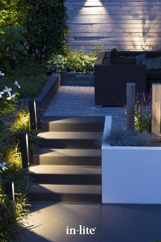in-lite ACE HIGH DARK 12v outdoor bollard lights, positioned upright in the borders alongside a garden staircase and patio levels, illuminating the borders, staircase and patio levels with a targeted light spread either side of the fixture. Low voltage outdoor garden lights, atmospheric path lighting for ambiance.