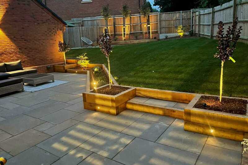 12 volt Up-Down wall light illuminating garden furniture in patio space and sleeper built raised planting beds with low voltage outdoor spotlights illuminating selected garden plants in client's unique outdoor space in Tidbury Green.