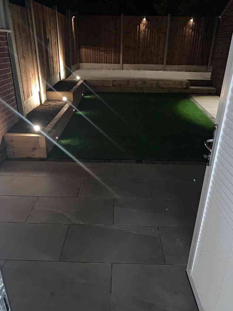 Low maintenance garden at night with outdoor porcelain, artificial lawn, raised patio terrace, new fencing and 12v garden lighting installed in Kenilworth, Warwickshire - Oakland Group.