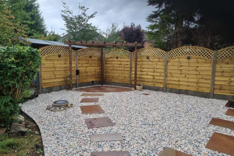 Decorative stone water feature, cobbles and gravel alongside natural stone pathway leading to natural stone patio area complete with pergola, curved trellis fencing panels and in-lite 12v outdoor wall lights mounted on fence panels.