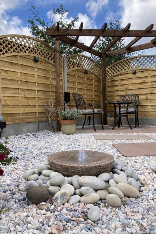 Decorative stone water feature, cobbles and gravel alongside natural stone pathway leading to natural stone patio seating area complete with pergola, curved trellis fencing panels and in-lite 12v outdoor wall lights mounted on fence panels.