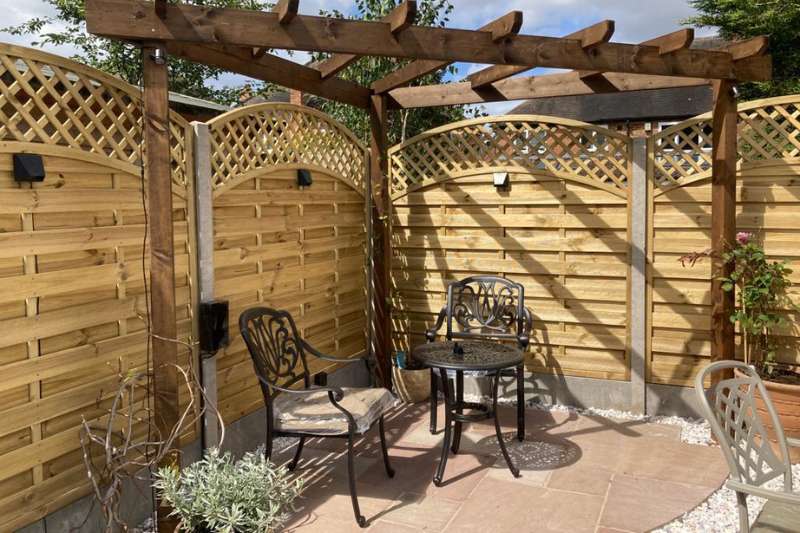 Natural stone patio area complete with garden furniture, pergola, curved trellis fencing panels and in-lite 12v outdoor wall lights mounted on fence panels.
