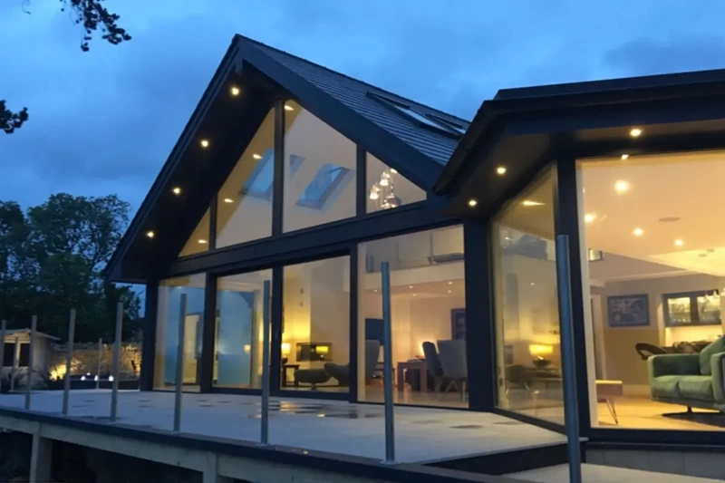 Large detached contemporary extension with exterior lights illuminating raised deck terrace, outdoor porcelain tiles and balustrade installed using the iGarden light steel subframe foundation system.