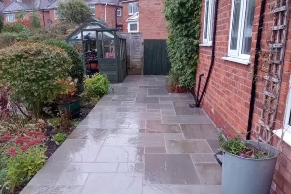 New patio and pathways built of natural stone paving slabs installed at the top of a sloping garden with planted borders and greenhouse area.